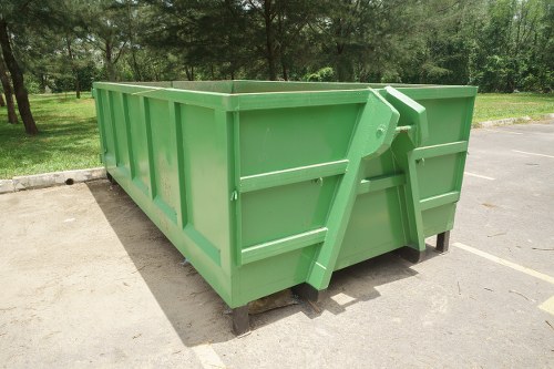 A large, empty green metal skip positioned on a paved surface in an outdoor setting, likely part of a rubbish removal service in Surbiton. The skip features vertical reinforced sides with visible ridges and a sturdy frame, and its bright green paint has a smooth, glossy finish. It has a hinged tailgate at the rear, secured with a locking mechanism, allowing for easy loading of waste materials. The skip rests on a black metal underframe, slightly elevated over the ground, with a wooden pallet visible underneath, providing stability on the paved area. In the background, there is a park or green space with trees and grass, suggesting the skip is situated in a suburban or urban environment typical of the Surbiton postcode area. The lighting is natural, indicating daytime, and the scene overall depicts a clean, well-maintained waste container ready for rubbish collection services, possibly coordinated by Commercial Waste Surbiton in the local area.