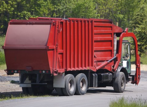 A large red refuse collection truck parked on a paved driveway or service road in a green, wooded area. The truck features a prominent rear waste hopper with vertical ridges, designed for collecting and compacting rubbish. Its textured metal surface shows signs of wear typical of waste management vehicles, with some reflective elements visible near the rear. The side of the truck includes a driver’s cab with a window, mirror, and hydraulic lifting mechanisms connected to the hopper, indicating it is used for rubbish collection and disposal. Surrounding the vehicle, there is grass and trees with fresh green foliage, suggesting a rural or suburban setting possibly in a town like Surbiton. The lighting appears natural, with daylight illuminating the scene, emphasizing the truck's bold red colour and the environment’s contrast. The scene aligns with rubbish removal services provided by companies such as Commercial Waste Surbiton, operating within the local postcode area, supporting efficient waste collection in the community.