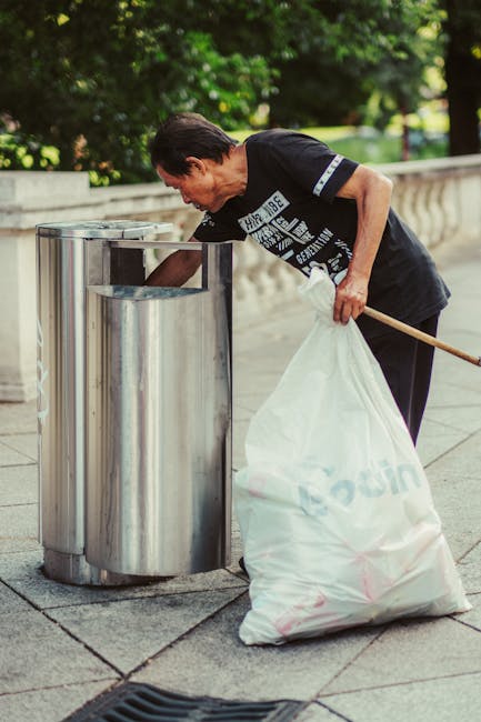 A black wheeled rubbish bin positioned on a suburban street at night, with the label 'ST. JOHN'S' on the front. The bin’s lid is open, revealing a pile of mixed waste including cardboard boxes, plas