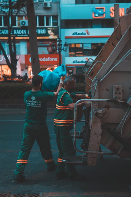 Two waste collection workers are engaged in a rubbish removal process on a city street in front of a large garbage truck. The worker on the left, wearing a dark green uniform with reflective orange an