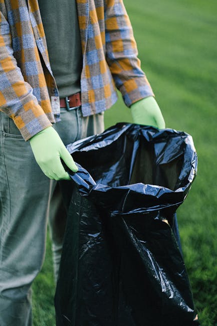 Two waste collection workers are engaged in a rubbish removal process on a city street in front of a large garbage truck. The worker on the left, wearing a dark green uniform with reflective orange an